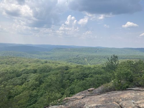 A panoramic view of lush green hills under a cloudy sky, seen from a rocky outcrop.