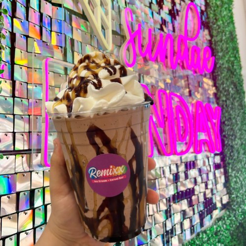 A hand holding a large chocolate sundae topped with whipped cream and drizzled with chocolate sauce against a colorful backdrop.