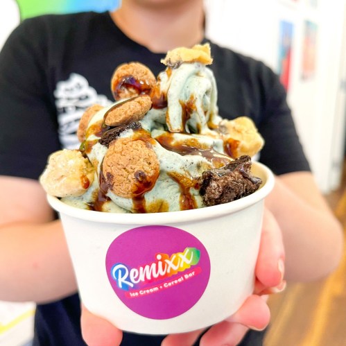 A person holds a cup of colorful ice cream topped with cookies, cereal, and chocolate drizzle.