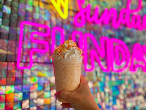 A hand holding a festive milkshake topped with whipped cream and sprinkles, in front of a colorful "Sundae Funday" sign.