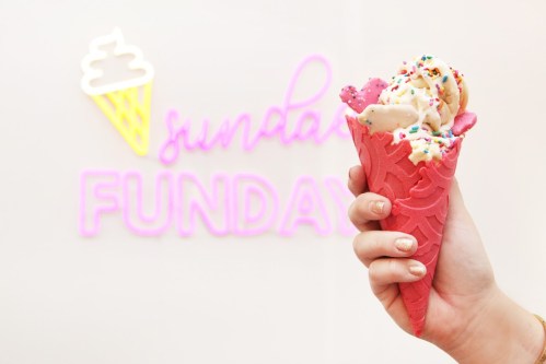 A hand holding a colorful ice cream cone in front of a "Sunday Funday" neon sign.