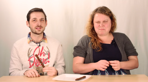 Two people sit at a table, one holding a chocolate-covered snack, while the other looks uncertain or displeased.