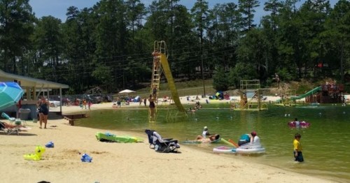 A sandy beach with people swimming, lounging, and playing near a lake, featuring slides and colorful floats.