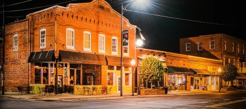 A charming brick building illuminated at night, with outdoor seating and nearby shops in a quaint town setting.