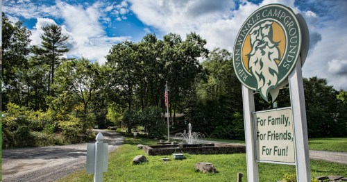 Sign for Ryan Winkle Campgrounds with a lion logo, surrounded by trees and a clear blue sky. Road leads into the camp.