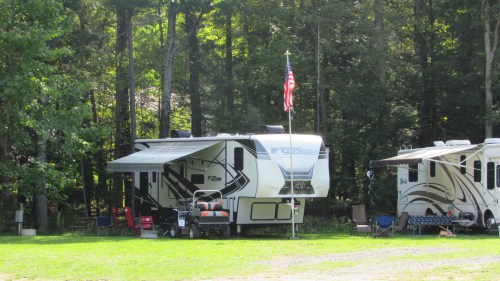 A parked RV with an American flag, surrounded by trees and outdoor seating in a grassy area.