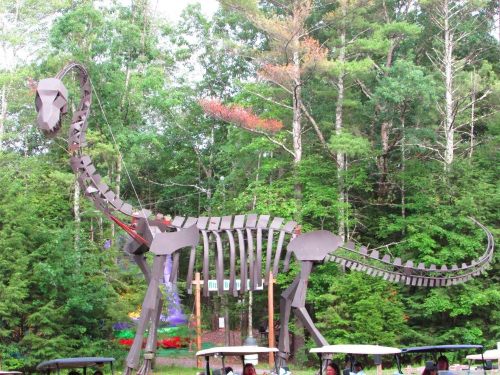 A large dinosaur skeleton sculpture stands among trees, with people and golf carts in the foreground.