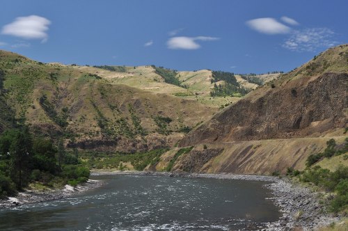 A winding river flows through a lush valley surrounded by rolling hills and a clear blue sky with scattered clouds.