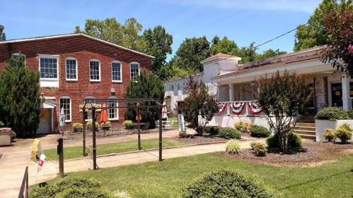 Historic brick buildings with greenery and a sunny sky, featuring a patio area and decorative flags.