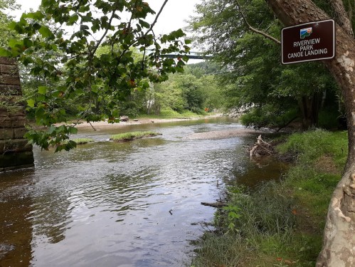 A serene river scene with a sign for Riverview Park Canoe Landing, surrounded by lush greenery and calm waters.