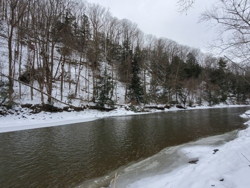 A serene river flows through a snowy landscape, bordered by bare trees and a snow-covered hillside.