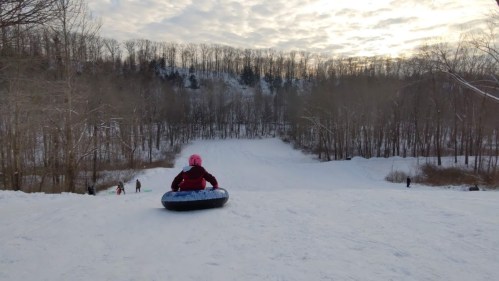 A child in a pink hat sits on a snow tube at the top of a snowy hill, ready to slide down. Trees line the background.