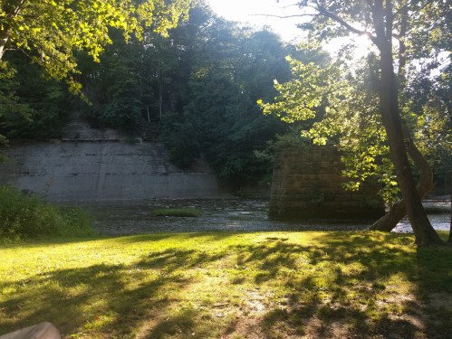 A serene riverside scene with lush greenery, a stone structure, and sunlight filtering through the trees.