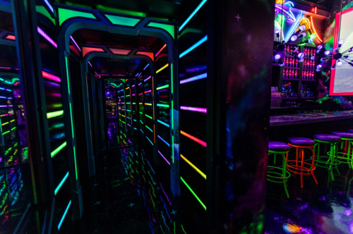 A vibrant hallway with neon lights in various colors, leading to a bar area with colorful stools and a cosmic theme.
