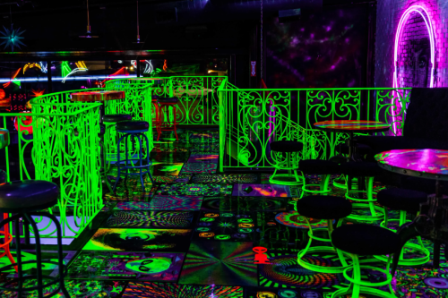 A vibrant, neon-lit bar with glowing green furniture and psychedelic patterns on the floor and walls.