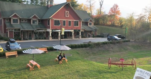 A cozy outdoor area with picnic tables, umbrellas, and a playground near a red building with shops. Trees in the background.