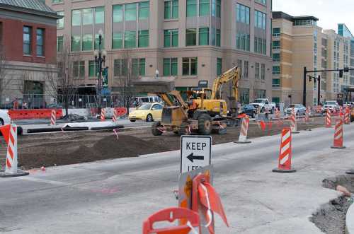 Construction site with heavy machinery, road barriers, and a "Keep Left" sign in an urban area.