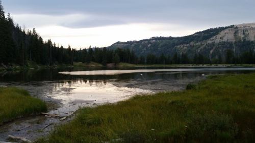 A serene lake surrounded by lush greenery and mountains, reflecting the cloudy sky at dusk.