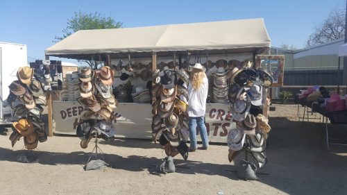 A woman browses a hat stall at an outdoor market, surrounded by various styles of hats hanging on displays.