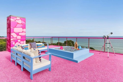 A vibrant rooftop terrace with pink carpet, blue furniture, a sandcastle, and a telescope overlooking the ocean.