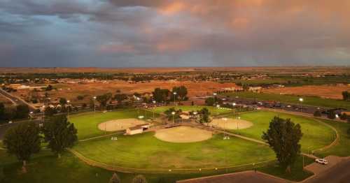 Aerial view of a baseball field at sunset, surrounded by trees and a small town in the background under cloudy skies.