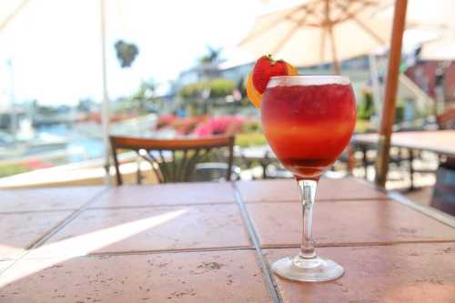 A colorful cocktail garnished with a strawberry and orange slice, set on a table with a scenic outdoor view.