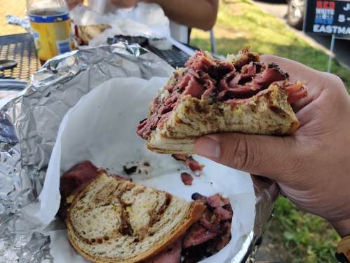 A hand holding a sandwich filled with layers of meat, with a partially eaten sandwich on a table in the background.