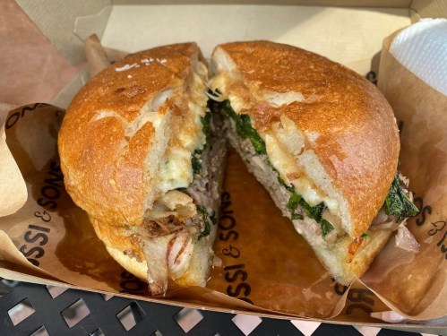 A freshly cut sandwich with meat, greens, and melted cheese, served in a takeout box lined with paper.