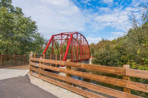 A red metal bridge spans a calm river, surrounded by trees and a wooden fence under a partly cloudy sky.