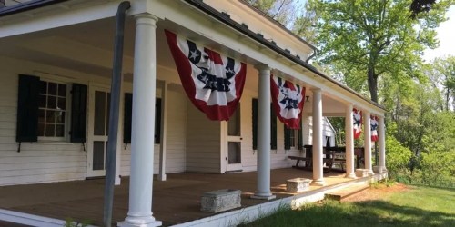 A white house with green shutters and patriotic bunting on the porch, surrounded by greenery on a sunny day.