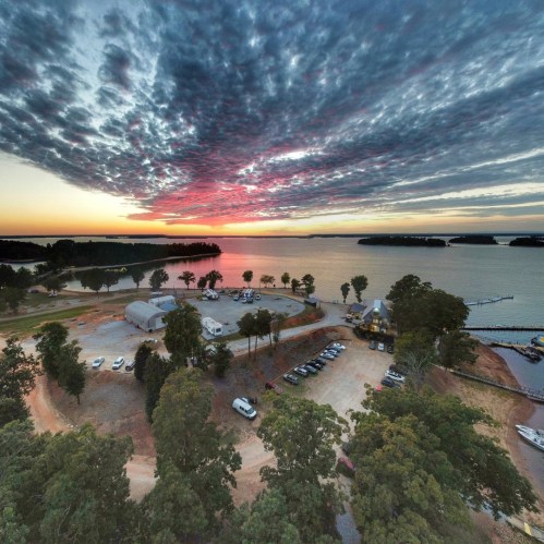 A scenic view of a lake at sunset, with colorful clouds and trees framing the shoreline and a parking area below.