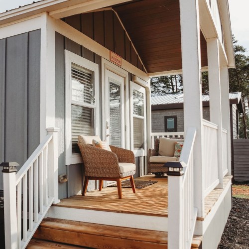 Cozy porch of a small house with a wicker chair and decorative pillows, surrounded by trees.