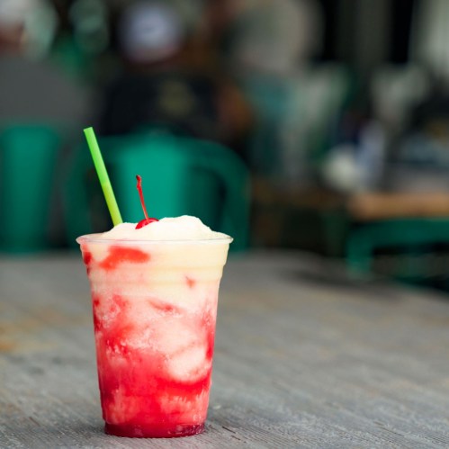 A colorful frozen drink in a clear cup with a green straw, featuring swirls of red and yellow.