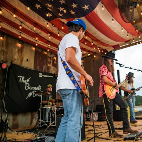 A band performs on stage with an American flag backdrop, featuring musicians with instruments and casual attire.
