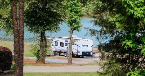 A camper parked near a lake, surrounded by trees and greenery, with a clear blue water view in the background.