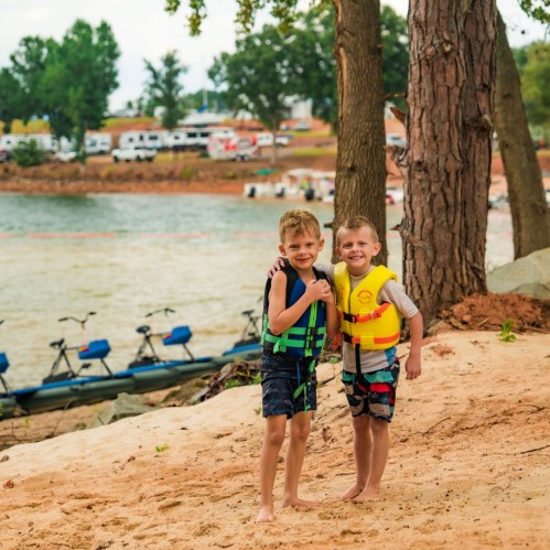 Two young boys in life jackets stand on a sandy shore by a lake, smiling and posing for the camera.