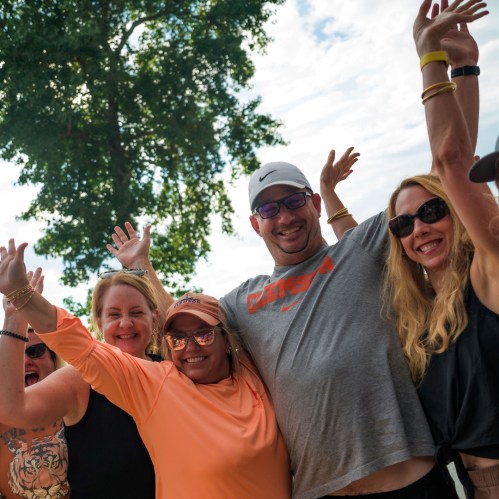 A group of smiling people celebrating outdoors with raised hands, surrounded by trees and a bright sky.