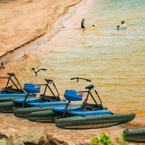 Three blue watercraft lined up on a sandy shore, with people swimming in the water nearby.