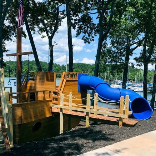 A playground featuring a wooden pirate ship structure with a blue slide, surrounded by trees and a marina in the background.