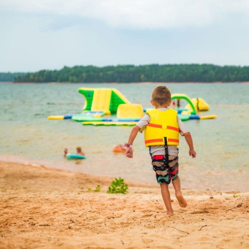 A child in a life jacket walks along a sandy beach, with inflatable water toys and people swimming in the background.