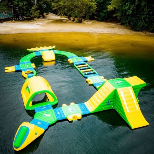 Colorful inflatable water park structure on a lake, featuring slides and climbing areas, surrounded by trees and sandy shore.