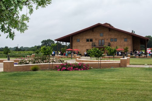 A rustic building surrounded by green grass and blooming flowers, with people gathered outside under red umbrellas.