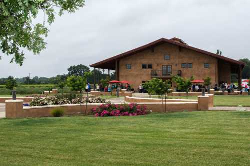 A rustic building surrounded by green grass and blooming flowers, with people gathered outside under red umbrellas.