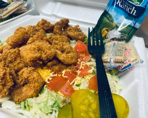 A takeout container with fried chicken on a bed of lettuce, tomatoes, cheese, pickles, and a side of ranch dressing.