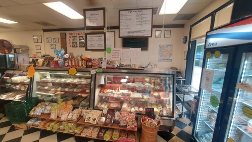 A deli display featuring sandwiches, desserts, and snacks, with a checkered floor and menu boards on the wall.