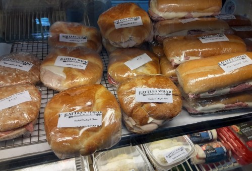 A display of various wrapped sandwiches in a deli case, labeled with names and prices.