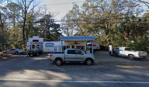 A small roadside shop with a blue awning, surrounded by trees and parked cars.