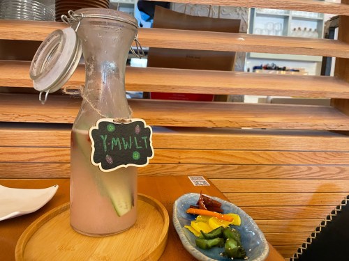 A glass bottle of drink with a label, accompanied by a small plate of colorful snacks on a wooden table.