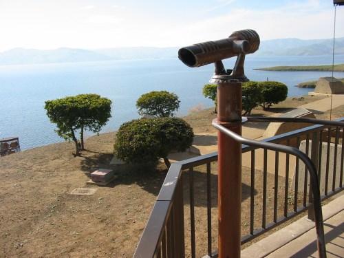 A telescope on a railing overlooking a calm lake with distant hills and green shrubs in the foreground.
