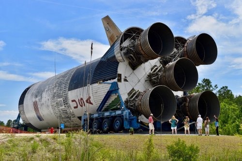 A large rocket on a transport vehicle, with people observing it in a grassy area under a blue sky.